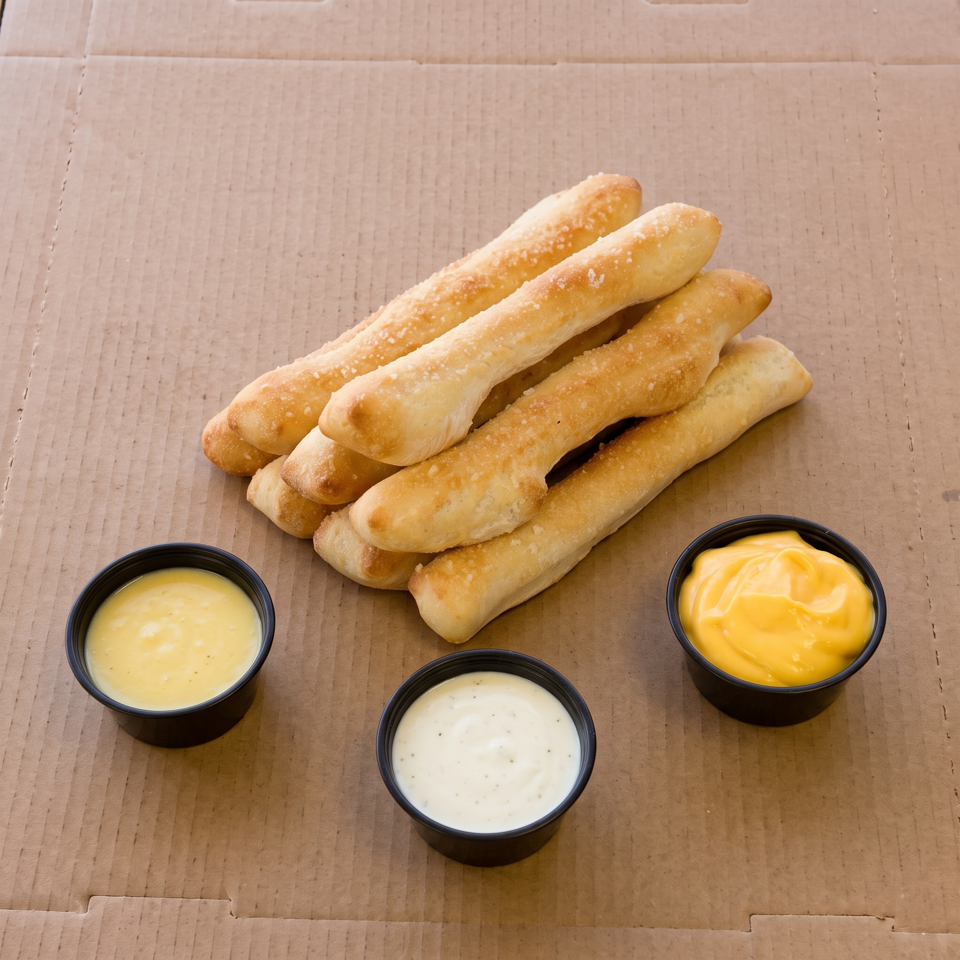 Golden-brown breadsticks with salt, served with three dipping sauces in black cups on a cardboard surface.